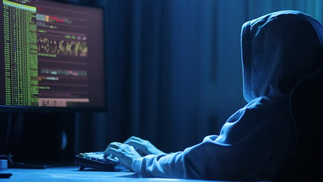 In a dimly lit room, a cloaked figure types code on a computer, hinting at a mysterious hacker