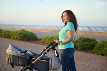 Curly haired multi ethnic brunette woman walking with her baby sleeping in pram, standing on the footpath on the promenade against Atlantic ocean on the background at sunset. Maternity leave lifestyle