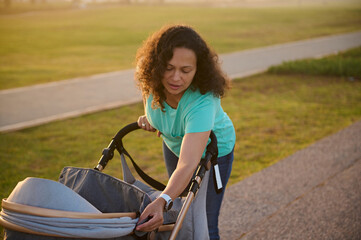 Obraz premium Curly haired happy mother looking and pushing infant baby stroller while walking in park. The concept of healthy active lifestyle. Daily outdoors walking and maternity leave