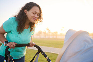 Obraz premium Positive smiling and happy young woman, loving mother looking t her newborn child while pushing the baby stroller in the city park at sunset. Mother taking care of her child. Copy advertising space