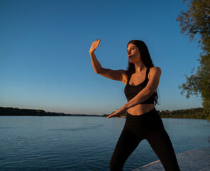 Young fit beautiful woman, practicing tai chi exercise by the river at sunset 