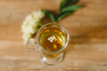 A clear glass mug filled with elderflower tea is placed on a rustic wooden table. Fresh elderflowers and a wooden cutting board are nearby, creating a cozy and natural atmosphere.