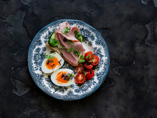 Delicious breakfast, snack - boiled egg, cherry tomatoes, cucumber and ham sandwich on a dark background, top view