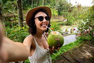 Young female tourist taking selfie with phone while drinking coconut water in Tegalalang rice...