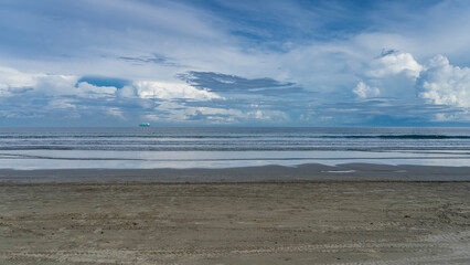 Morning on the ocean coast. Turquoise waves calmly roll towards the shore, spreading along the beach. There are tracks of cleaning machines in the sand. A ship is on the horizon. Clouds, blue sky