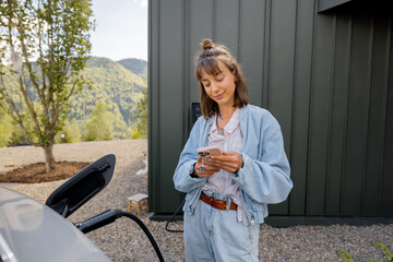 Young woman plugs a charger into electric vehicle standing with smart phone near her house...