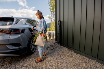 Young woman plugging a charger into electric vehicle near her house outdoors. Concept of modern...