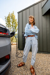 Portrait of a young woman plugging a charger into electric vehicle near her house outdoors. Concept...
