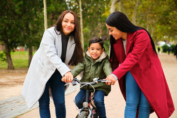 Latin lesbian couple helping their son to ride a bicycle in a park. LGBT family.