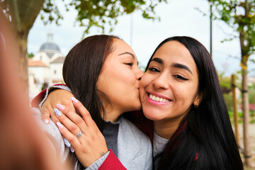 Happy Hispanic lesbian couple kissing and taking a selfie at street. LGBT couple.