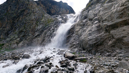 A powerful waterfall cascades down a rugged, rocky mountain face, surrounded by steep cliffs and boulders. The water rushes over the rocks, creating a misty spray at the base.