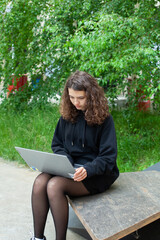 Photo with a portrait of a beautiful teenage girl with curly hair uses a laptop in the city on the street. 
