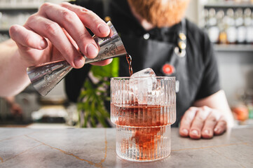 close-up person pouring pink beverage from metal jigger into textured glass over ice cubes. focus...