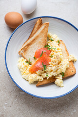 Toasts with scrambled eggs and smoked salmon served in a blue and white plate, vertical shot on a beige stone background