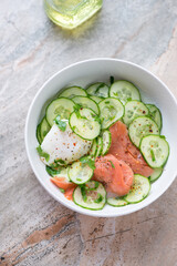 Cucumber salad with smoked salmon and poached egg in a white bowl, vertical shot on a grey and roseate granite background, elevated view