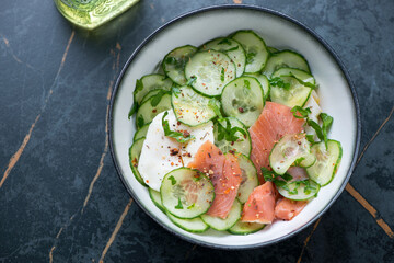 Bowl with smoked salmon and cucumber salad on a dark-green marble background, horizontal shot, high angle view