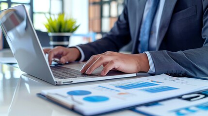 Businessman typing on a laptop with financial charts and graphs on a desk, analyzing data in an office setting.