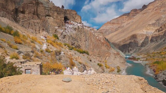 Wide angle shot of Phugtal monastery besides the blue Tsarap Chu river flowing besides the 2500 year old monastery built on a cave at Zanskar Valley in remote area of Ladakh, India. Birds fly in sky.
