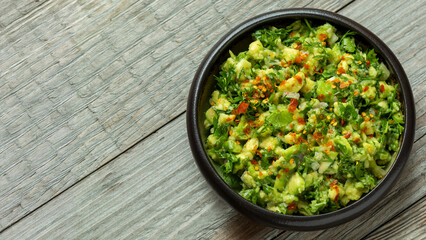 A bowl of homemade chunky guacamole with a lot of chopped fresh coriander, topped with hot chili flakes. Rustic wooden table. Baner with space for text.