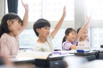 逆光の綺麗な明るい教室で授業をうけ勉強する元気な小学生 ハイと手を挙げ挙手をする