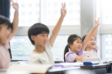 逆光の綺麗な明るい教室で授業をうけ勉強する元気な小学生 ハイと手を挙げ挙手をする