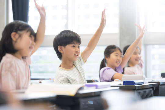 逆光の綺麗な明るい教室で授業をうけ勉強する元気な小学生 ハイと手を挙げ挙手をする
