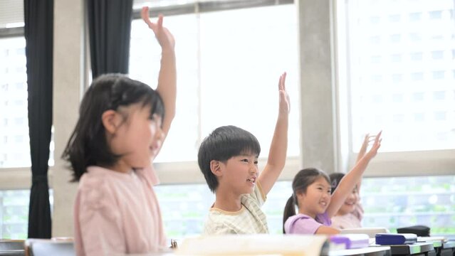 逆光の綺麗な明るい教室で授業をうけ勉強する元気な小学生 ハイと手を挙げ挙手をする