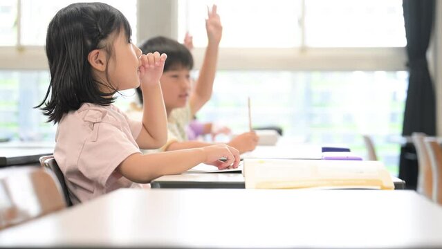 逆光の綺麗な明るい教室で授業をうけ勉強する元気な小学生 ハイと手を挙げ挙手をする