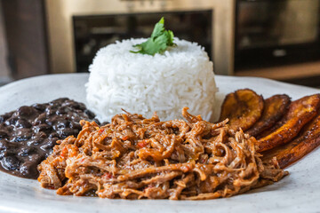 Plato tradicional venezolano de pabellón criollo con carne mechada, arroz, caraotas negras y plátanos fritos. En un entorno de cocina con iluminación natural, evocando un ambiente casero y cálido.