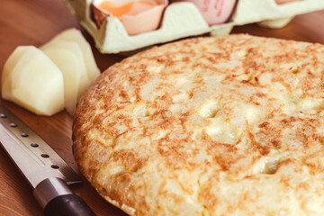 Una tortilla de patatas dorada, acompañada de huevos y patatas frescas, sobre una tabla de madera en una cocina bien iluminada. El ángulo cercano destaca la textura esponjosa y la corteza dorada.