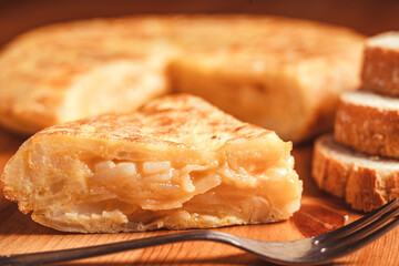 Primer plano de una deliciosa tortilla de patatas española, con una textura jugosa y acompañada de rodajas de pan rústico. Un tenedor de metal se encuentra en primer plano sobre la mesa de madera.