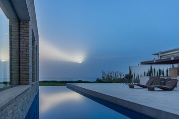 A modern villa with an infinity pool overlooking the ocean at dusk. The sun is setting over the...