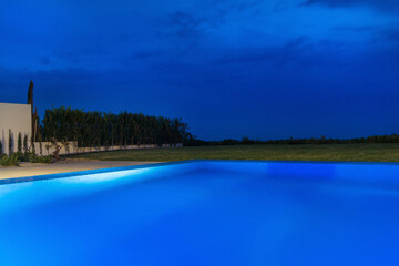 Nighttime View of a Swimming Pool With a Blue Sky and Grassy Area. The pool is illuminated with blue lighting.