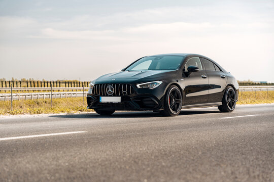 Photo of a black Mercedes-Benz AMG CLA 45 S 4MATIC+. They show details of the car from the outside. Rims, emblems, headlights, lamps in a sporty style. The model stands in the middle of highway.