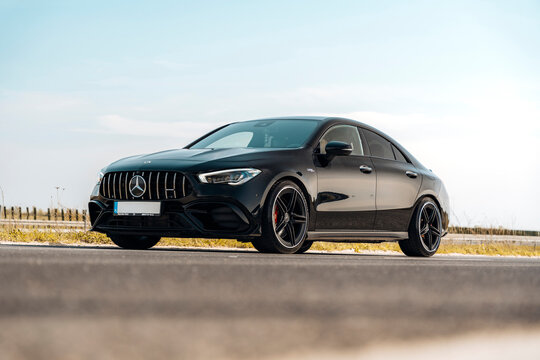 Photo of a black Mercedes-Benz AMG CLA 45 S 4MATIC+. They show details of the car from the outside. Rims, emblems, headlights, lamps in a sporty style. The model stands in the middle of highway.
