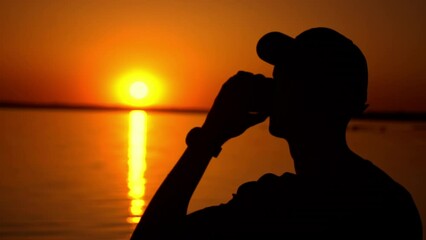A man stands in silhouette, sipping from a cup as he gazes out over the tranquil lake at sunset. The vibrant orange sky creates a breathtaking backdrop for a moment of peaceful reflection.