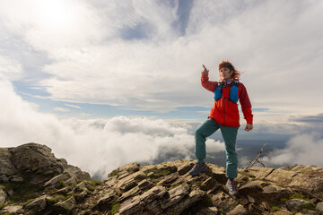A woman in a red jacket stands on a rocky mountain top, pointing to the sky