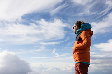 A man in an orange jacket stands on a mountain top, looking out at the sky