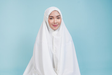 Smiling young Asian Muslim woman wearing prayer gown praying while sitting down over isolated by blue background. Hajj concept.