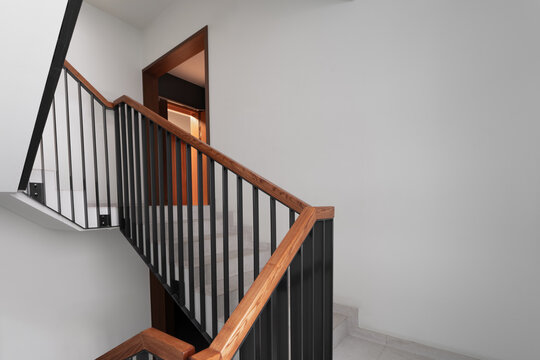 A contemporary staircase design featuring a sleek wooden handrail and black metal balusters, leading up to a well-lit hallway with minimalistic decor and clean lines. - Powered by Adobe