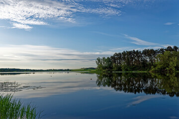 Dawn at Rescobie Loch Fishery near to Forfar, with the reflections of the wooded banks opposite in the calm blue water beneath a blue sky.