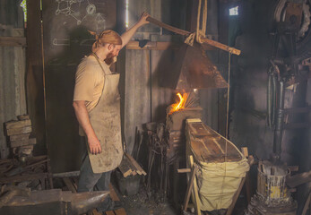 Blacksmith in an authentic forge fans the fire with hand-held bellows. Traditional crafts