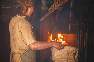 Blacksmith in an authentic forge fans the fire with hand-held bellows. Traditional crafts