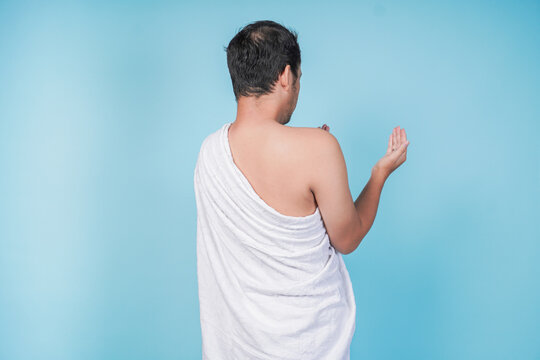 Back portrait of young Asian Muslim man wearing ihram praying with hand gesture over isolated by blue background. Hajj concept.