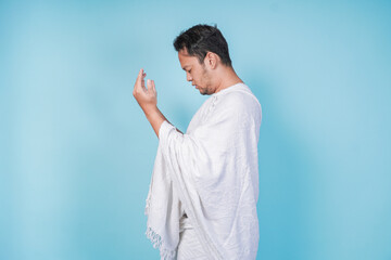 Side portrait of young Asian Muslim man wearing ihram praying with hand gesture over isolated by...