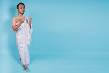 Smiling young Asian Muslim man wearing ihram praying while walking over isolated by blue background. Hajj concept.