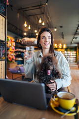 Beautiful and happy young woman sitting in modern pet shop cafe bar and enjoying in fresh coffee together with her adorable brown toy poodle.