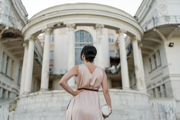 A woman in a pink dress stands in front of a large building. She is looking at the camera with a serious expression. The building is old and has a grand appearance. The woman's dress is elegant.
