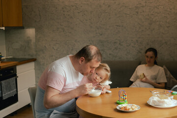 Close-up of a father with his daughter in his arms feeding her. Mom is busy with kitchen chores. The concept of genderblend and role reversal in the family. Part of the series