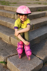 A small and cheerful girl in a pink helmet and roller skates is sitting on the stairs